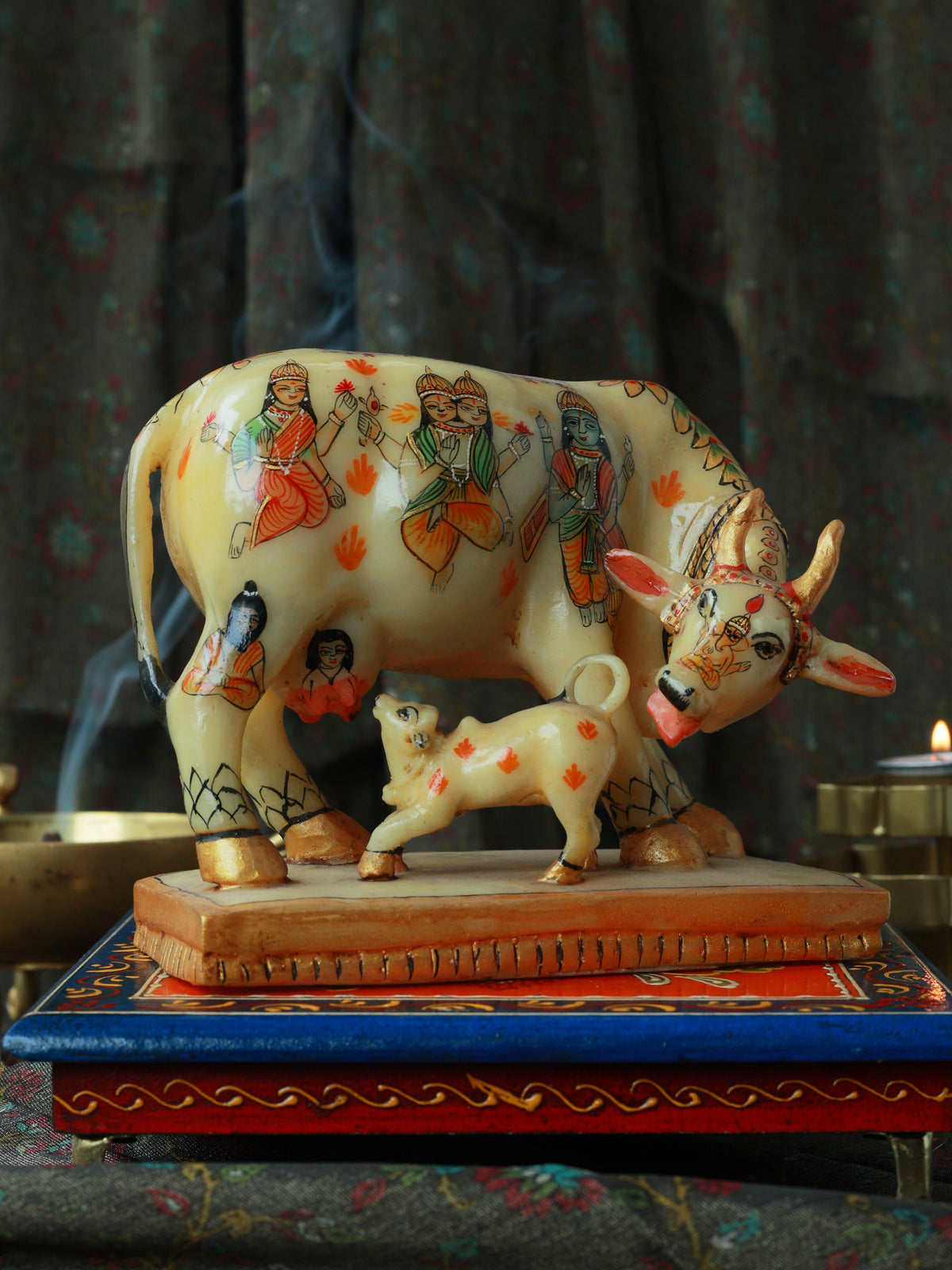 A hand-painted clay cow and calf figurine decorated with traditional Indian motifs, resting on a decorative blue and red tray against a dark, smoky background.