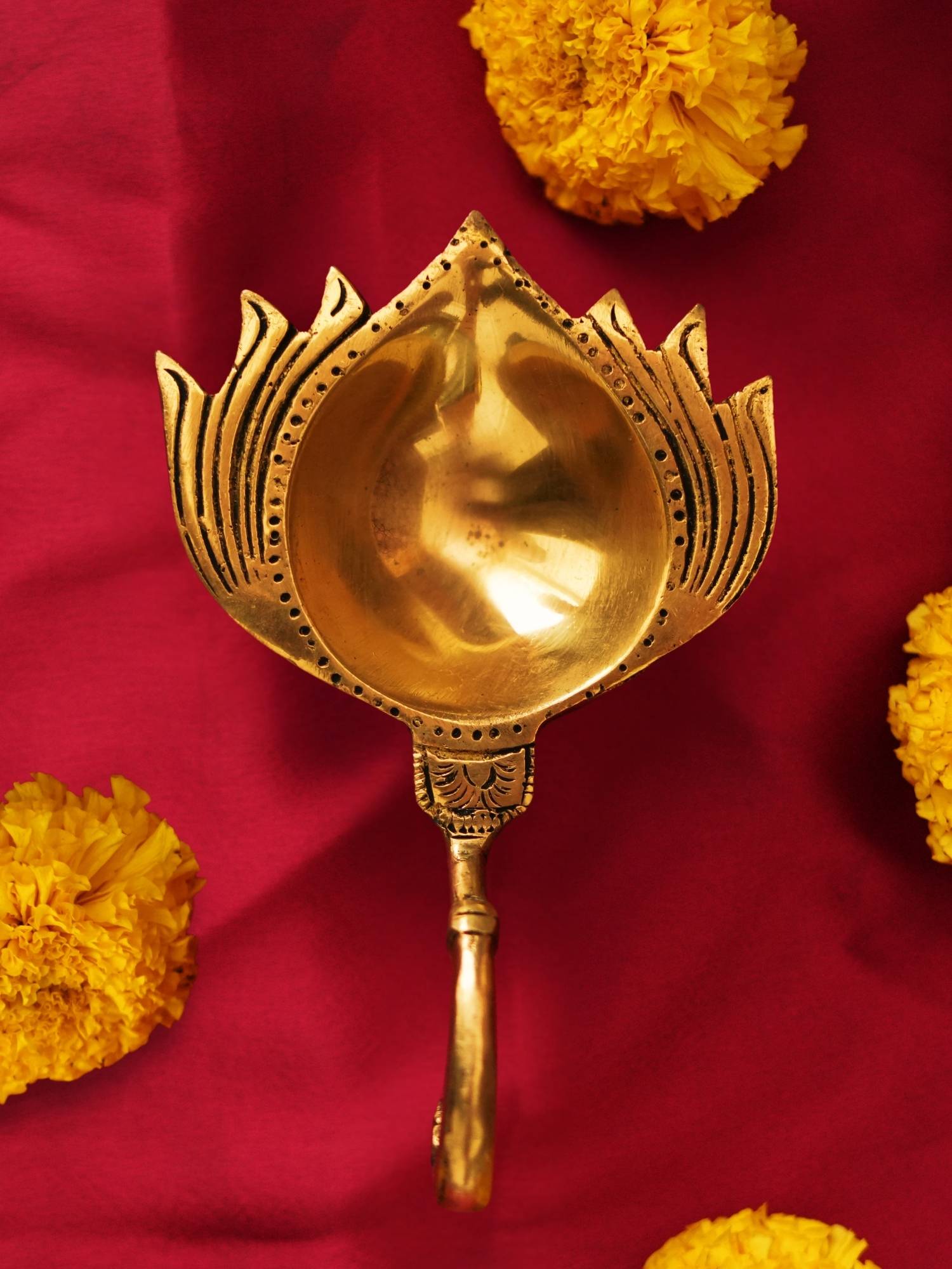 A brass lotus-shaped diya with intricate designs and a handle, displayed against a red background with yellow flowers scattered around.
