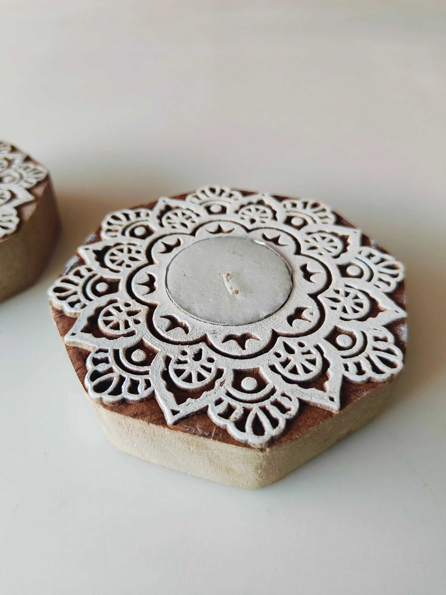 A handmade wooden block tealight holder with a round shape and a white mandala-style pattern, containing a lit tealight candle.