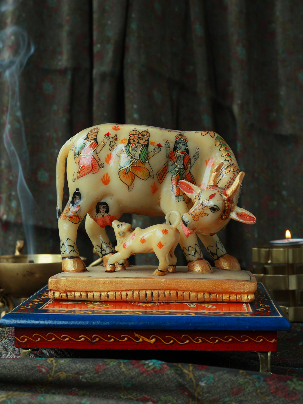 A decorative hand-painted cow and calf figurine displayed on a colorful ornate stand, with a smoky incense burner and candle in the background.