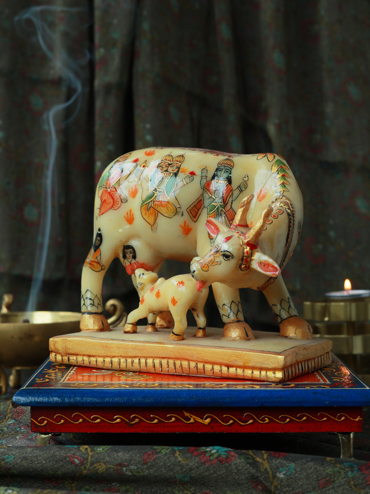 A decorative painted cow and calf figurine standing on a rectangular base, displayed on a blue and red decorative stand against a dark patterned curtain background with a smoky incense burner and candle visible.