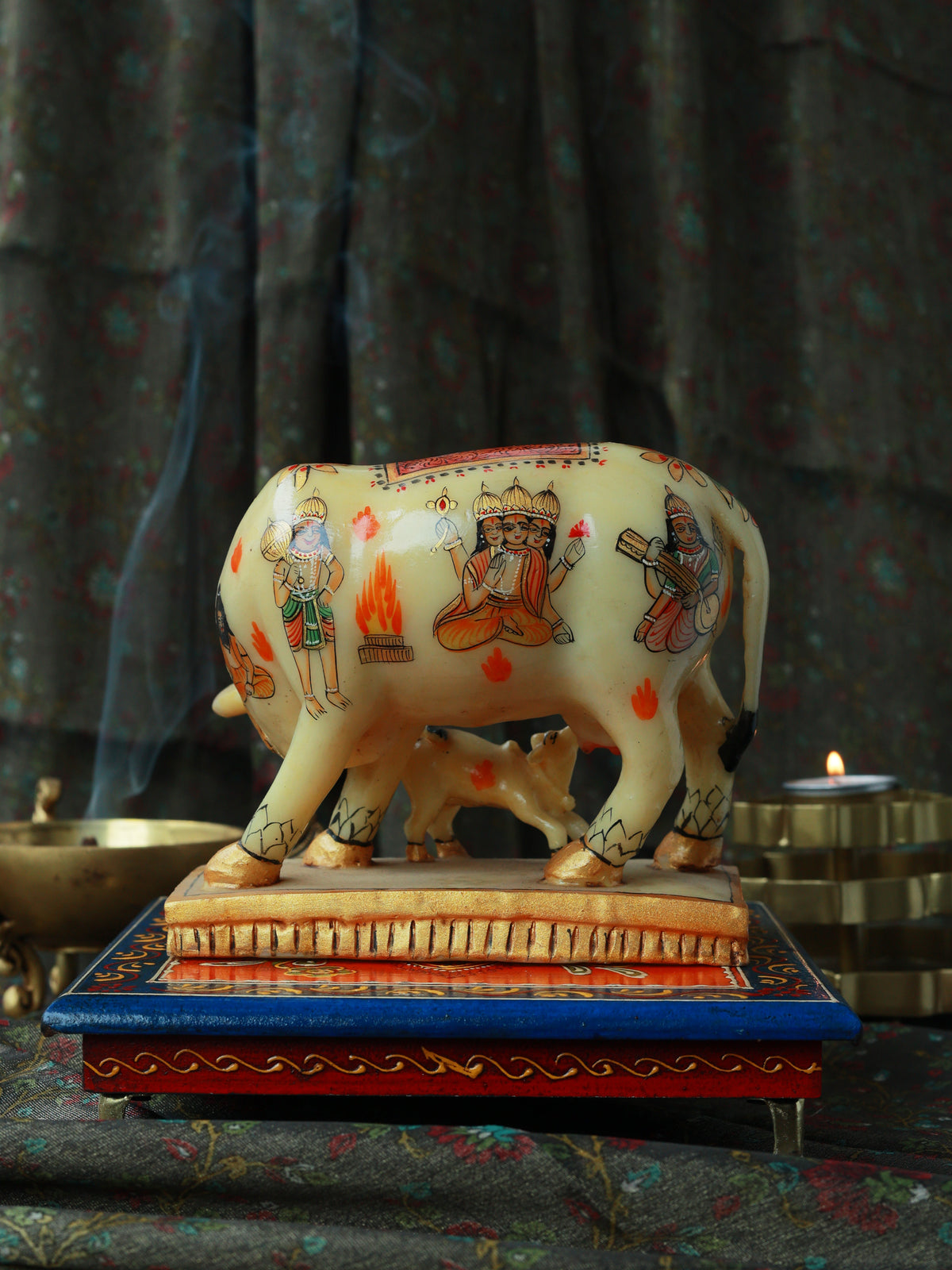 A decorative cream-colored cow and calf figurine with hand-painted traditional motifs, displayed on a blue and red patterned base against a dark draped background with subtle smoke and candlelight.