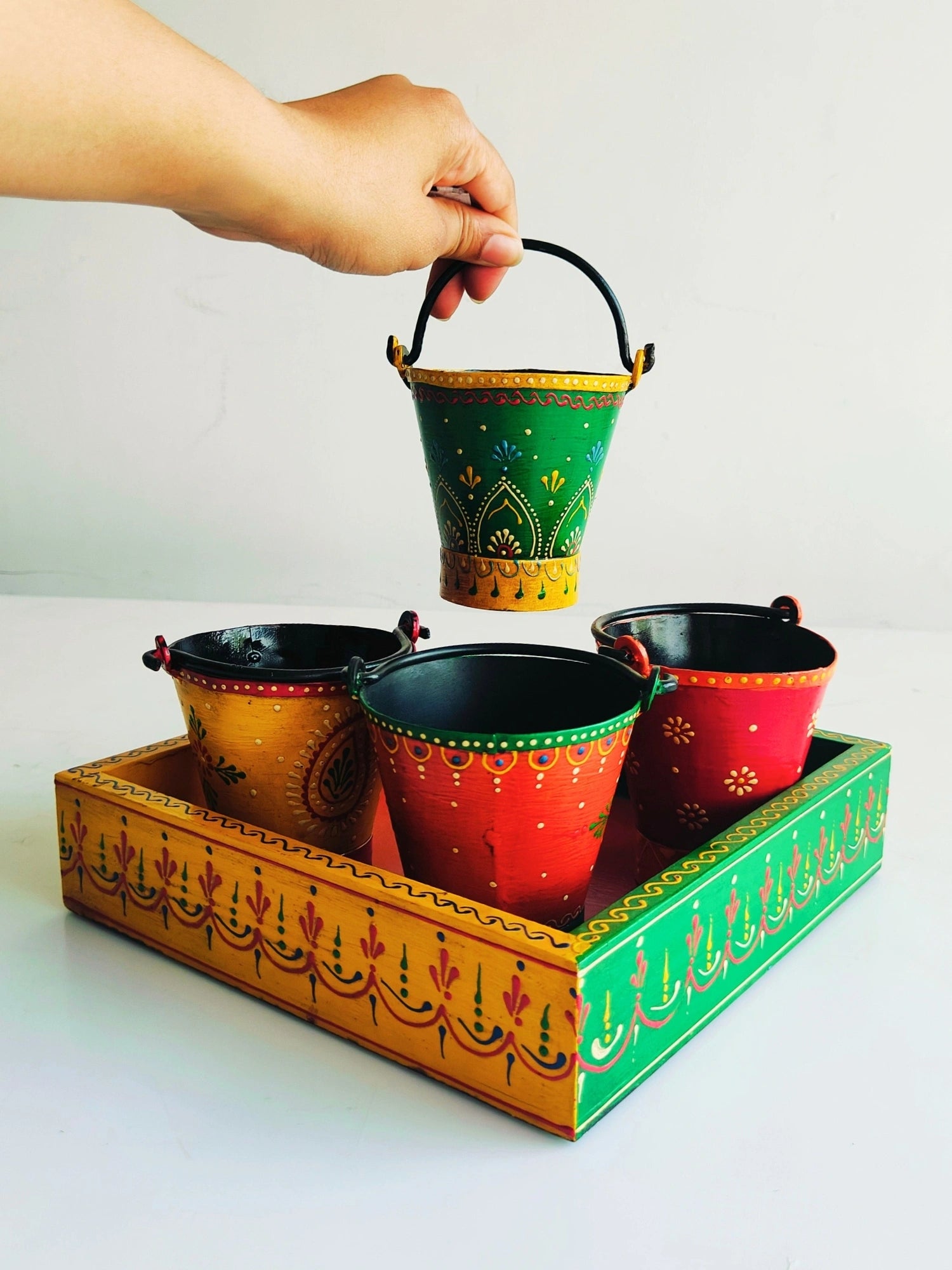 A hand lifting a colorful hand-painted bucket with a handle, out of a vibrant hand-painted wooden platter, which is placed on a white surface.